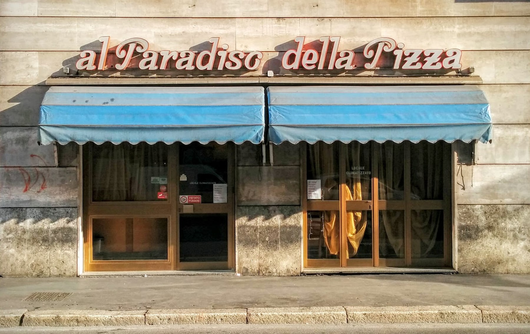La facciata della pizzeria con l'insegna in corsivo e le tende a righe azzurre