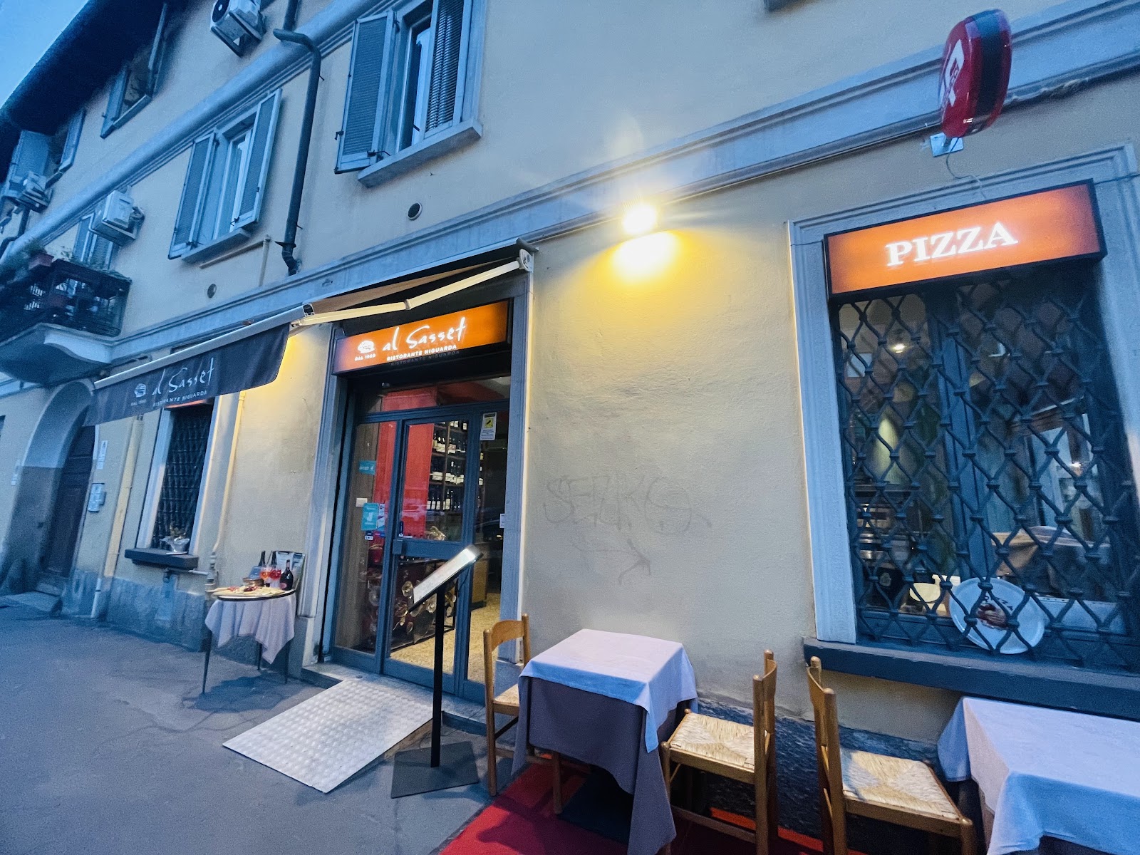 L'ingresso di al Sasset in Via Graziano 35 all'imbrunire, con l'insegna arancione e i tavolini apparecchiati fuori