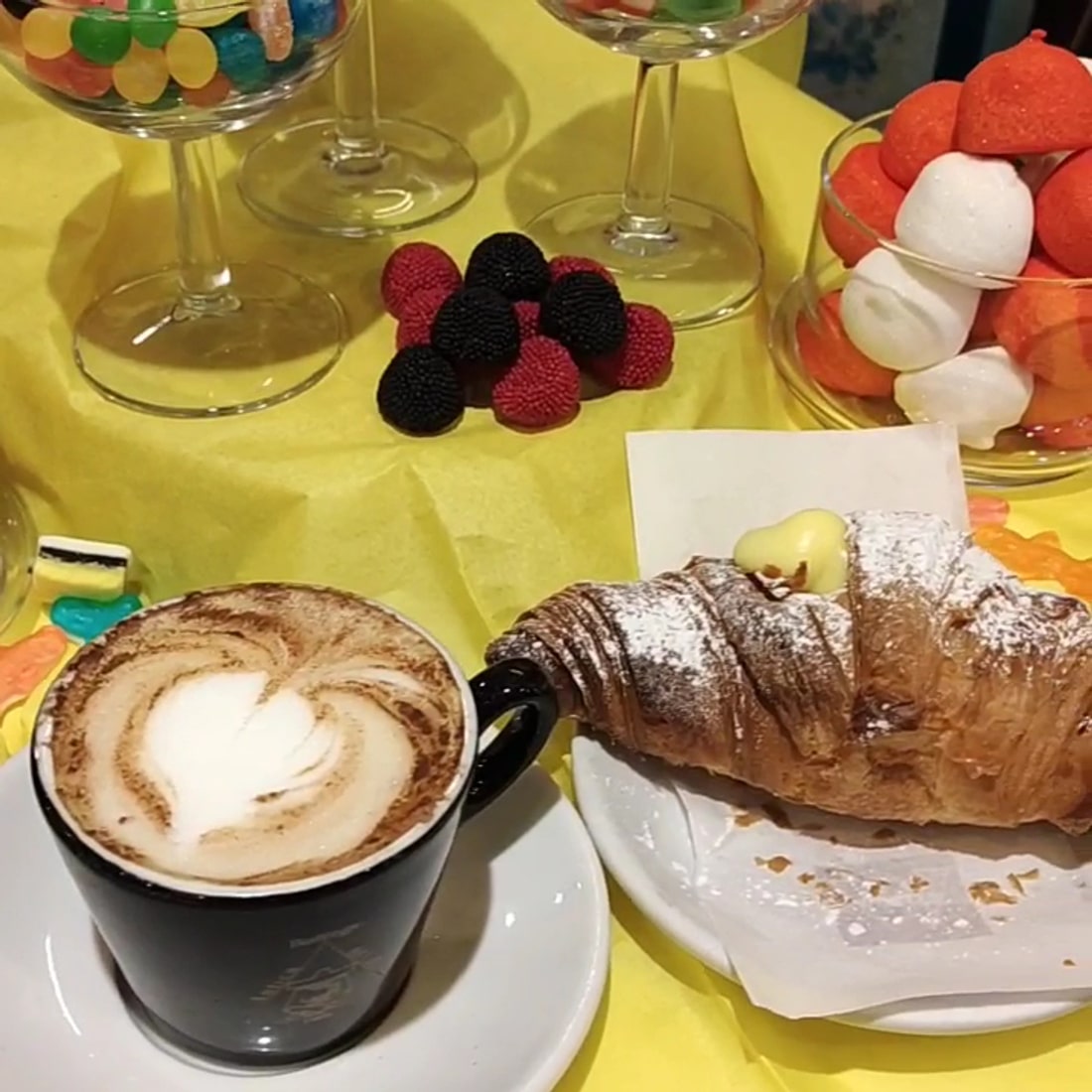Un cappuccino con latte art a cuore e un cornetto alla crema, con le caramelle nelle coppe di vetro sullo sfondo