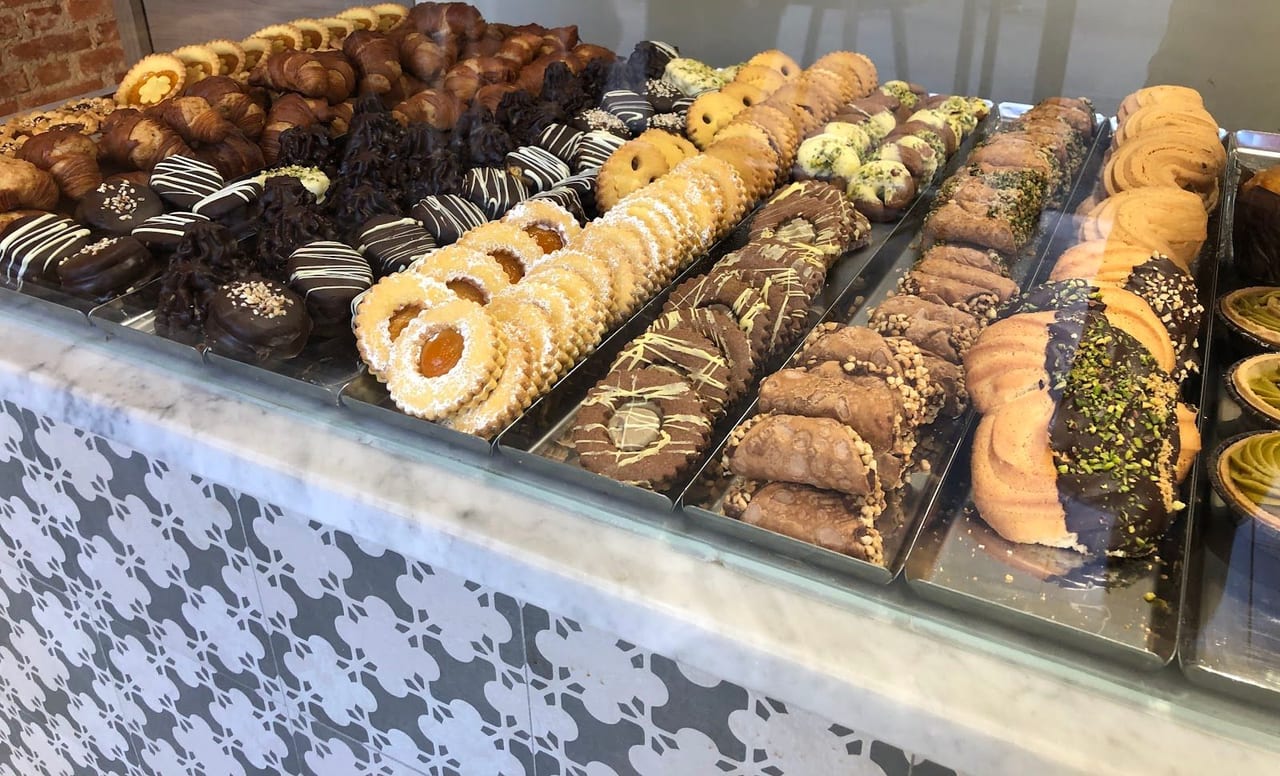 Il banco di marmo con le cementine grigie e la pasticceria secca: occhi di bue, cannoli, frolle al cioccolato e al pistacchio.