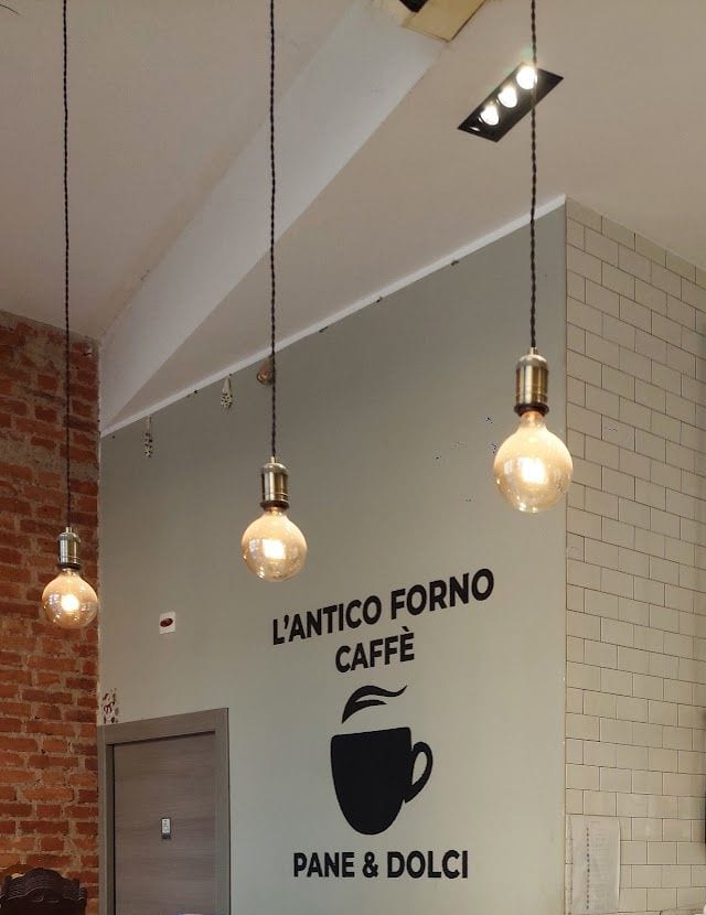 Il logo nero sul muro grigio: «L'Antico Forno, Caffè», la tazzina, «Pane & Dolci»; le lampadine e le piastrelle bianche.