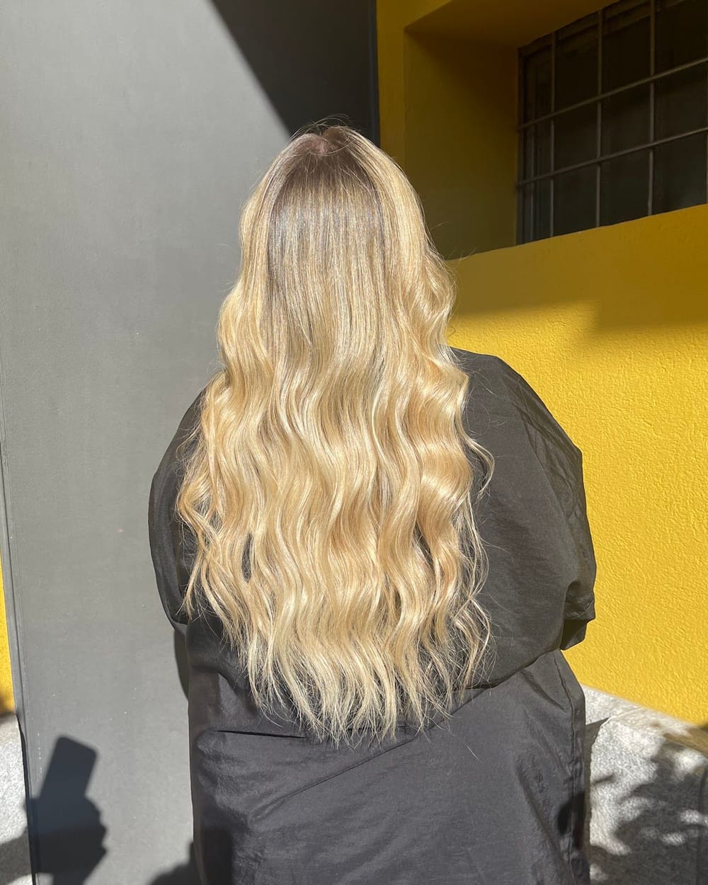 Capelli lunghi e mossi, biondi, con la radice scura e sfumata, di spalle davanti a un muro giallo