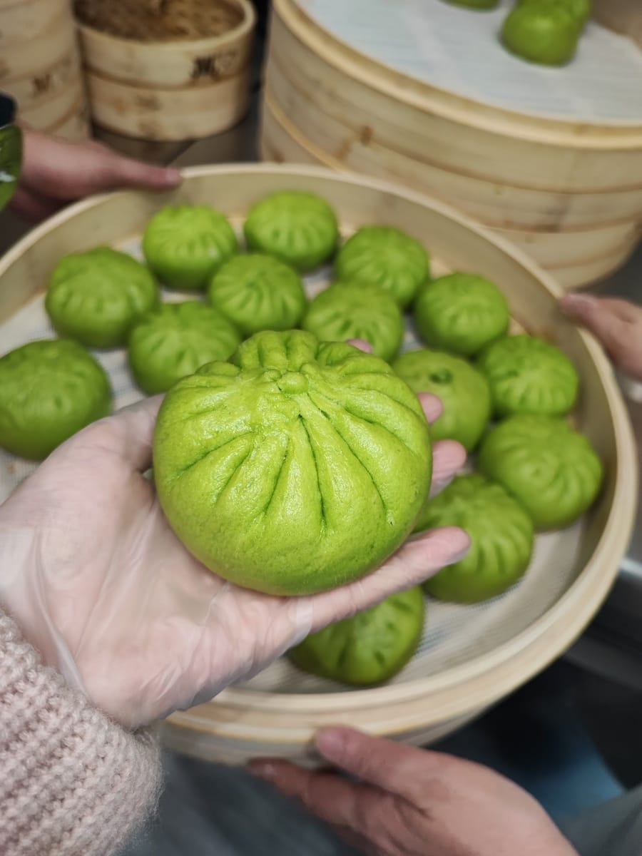 Una vaporiera piena di bao verdi, uno sollevato con la mano guantata.