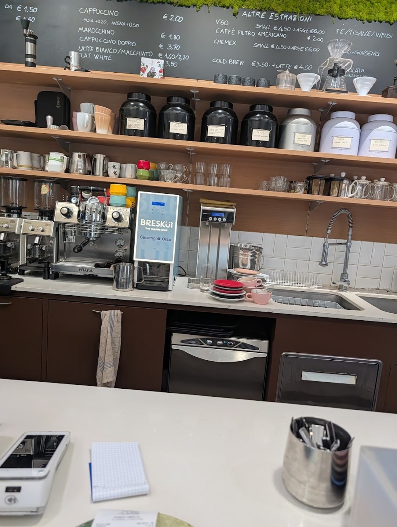 Il banco con due macchine espresso, i V60 e la Chemex sullo scaffale e il menù delle estrazioni scritto sulla lavagna a muro