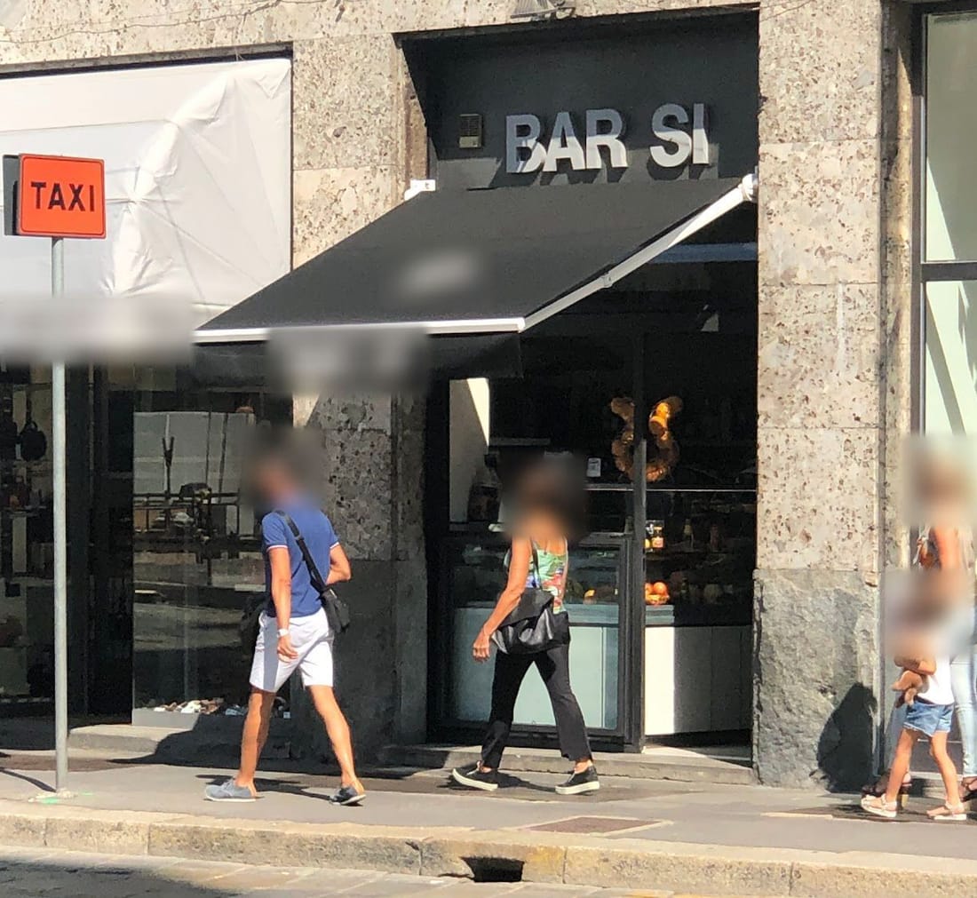 La tenda nera con BAR SI sopra la vetrina, all'angolo, e il cartello arancione del taxi davanti.