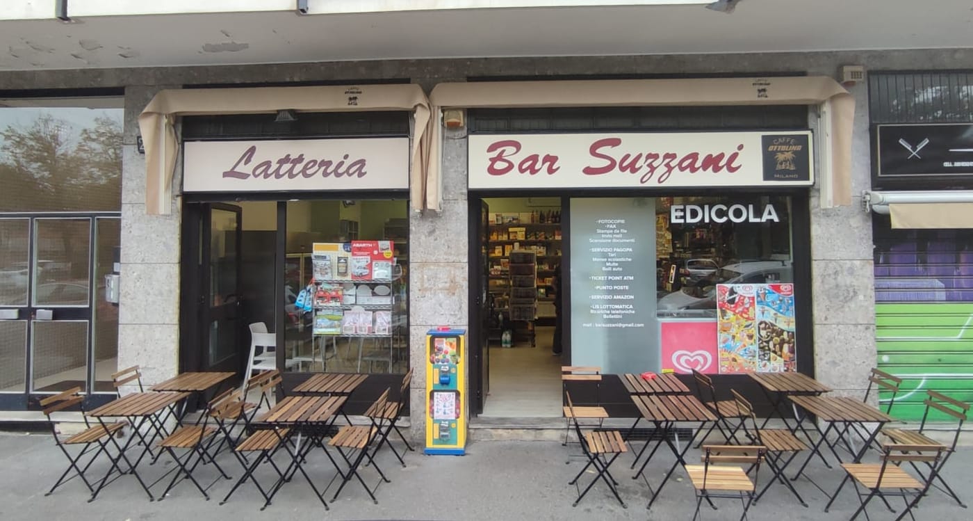 La facciata del Bar Suzzani con le tre insegne Latteria, Bar Suzzani ed Edicola e i tavolini sul marciapiede