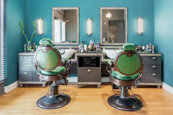 La sala del taglio di Barbieria 88: pareti verde petrolio, due poltrone da barbiere in pelle verde smeraldo, specchi con cornice geometrica e applique scanalate