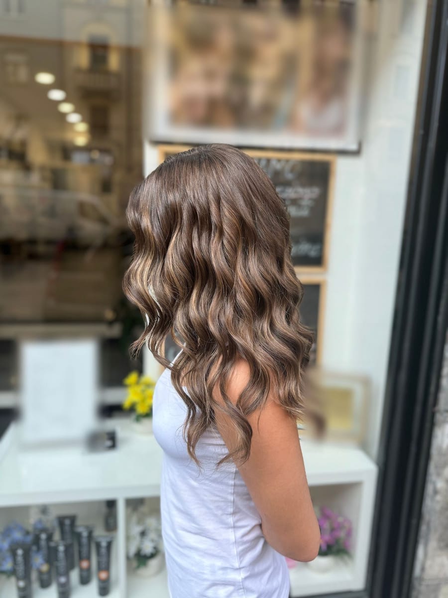 Capelli lunghi castani con le onde, di spalle, alla vetrina del salone.