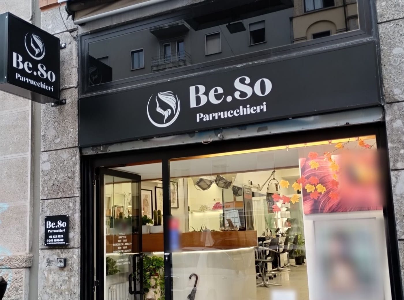 La vetrina di Be.So Parrucchieri: l'insegna nera con il nome in bianco, la bandiera, la targhetta coi numeri e l'adesivo degli orari sulla porta.