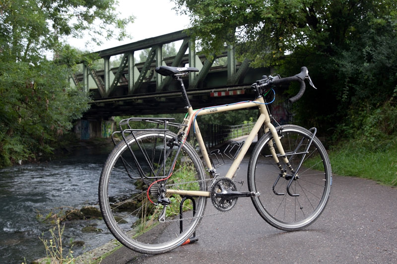 Bici da viaggio color crema appoggiata a un muretto lungo un fiume, con un ponte di ferro alle spalle