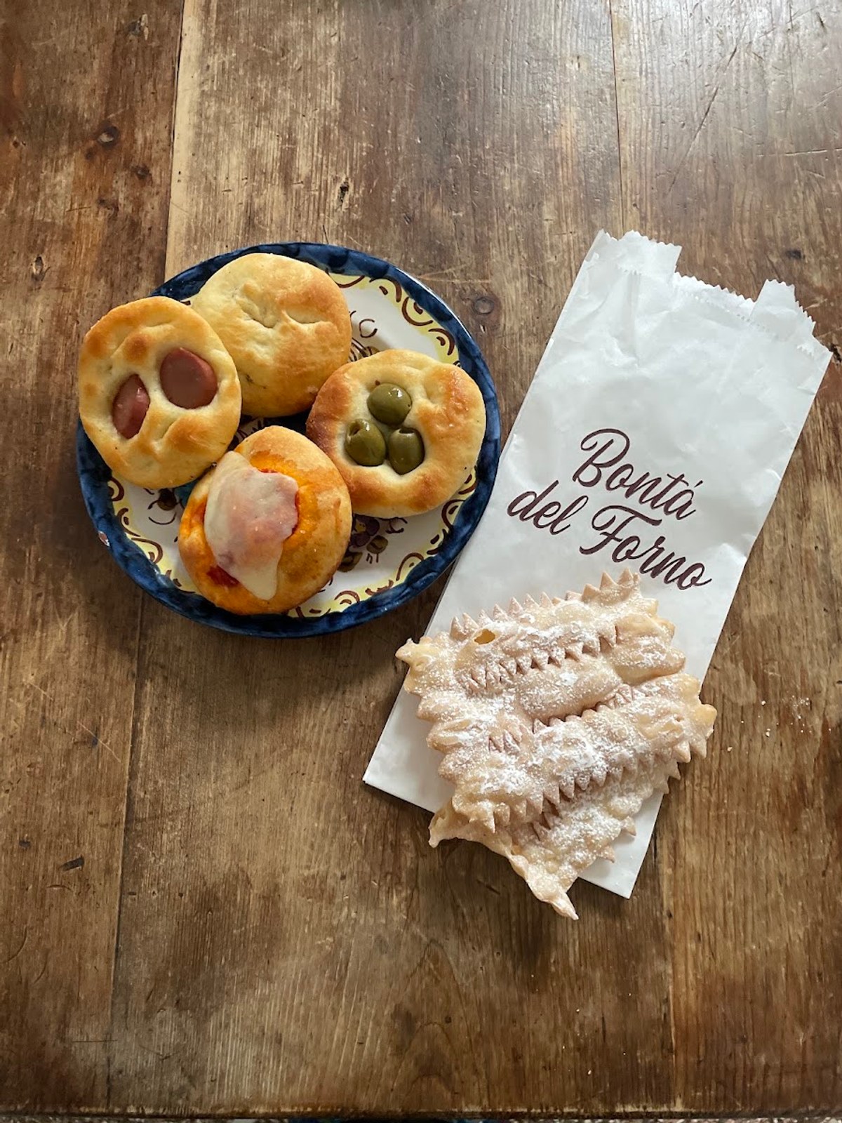 Un sacchetto di carta con «Bontà del Forno» scritto in corsivo, accanto a pizzette con würstel e olive e a delle chiacchiere
