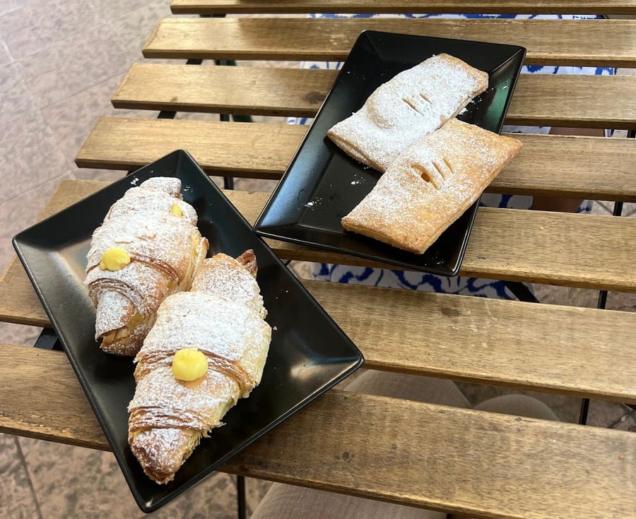 La colazione su un tavolino di legno: due cornetti alla crema e due sfoglie su piatti neri.
