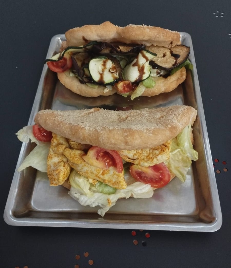 Due focaccine baciate farcite sul vassoio d'alluminio: una con le verdure grigliate e la glassa scura, una con straccetti di carne, insalata e pomodoro.