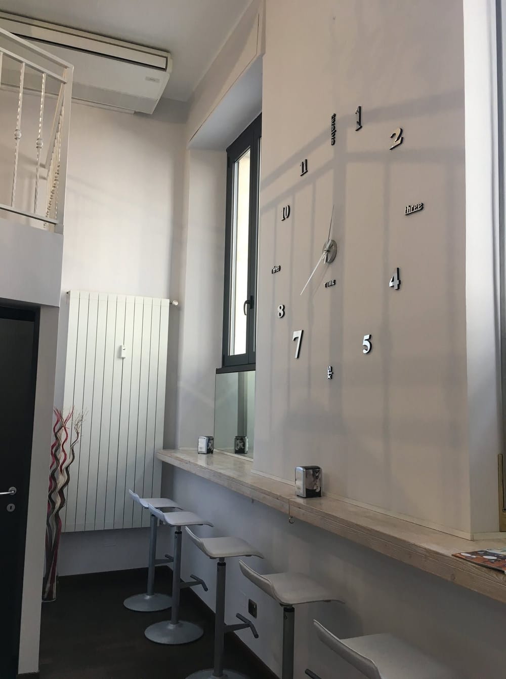 Il banco lungo la parete con gli sgabelli bianchi e, sul muro, l'orologio coi numeri sparsi.