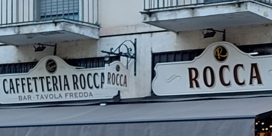La facciata in Via Binda: i due cartigli crema con Caffetteria Rocca e Rocca, il disco con la R d'oro, l'insegna a bandiera sul braccio di ferro battuto e la tenda grigia