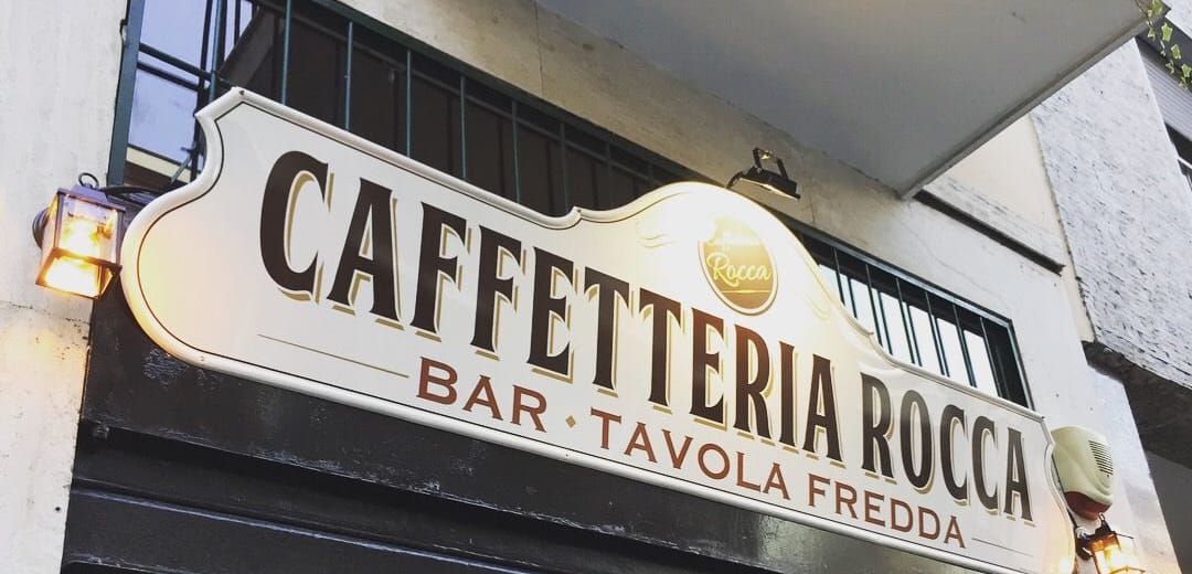 L'insegna crema a cartiglio con la scritta Caffetteria Rocca e, sotto, Bar · Tavola fredda, il disco d'oro col marchio e una lanterna accesa