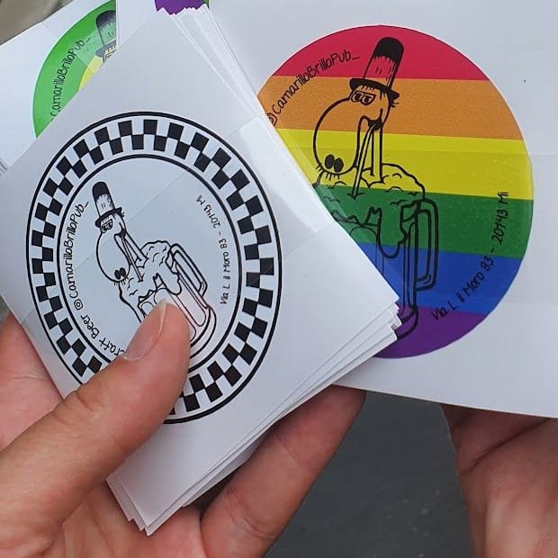 Due mani che tengono i loro adesivi tondi: il personaggio col cilindro che beve dal boccale, in versione a scacchi e arcobaleno, con l'indirizzo del pub