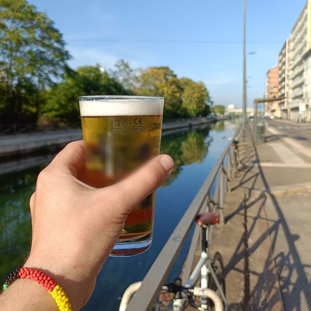 Una birra tenuta in mano sopra l'acqua del Naviglio, con il parapetto e una bicicletta