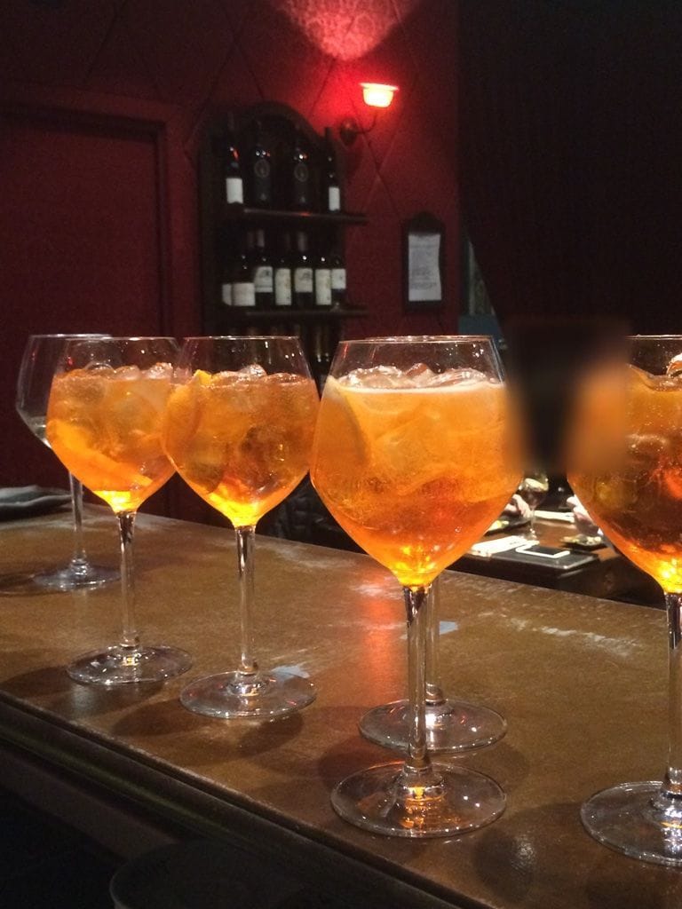 Gli spritz in fila sul bancone, dietro la parete rossa con la lampada.