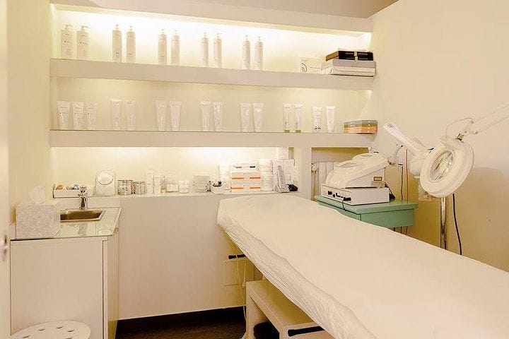 Una cabina bianca: il lettino, la lampada con la lente, le mensole illuminate coi flaconi.