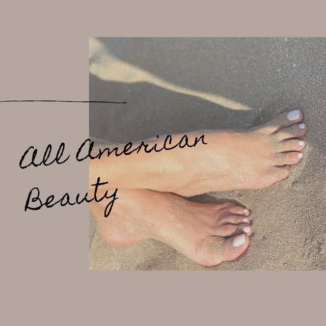 I piedi sulla sabbia con lo smalto chiaro, e la scritta All American Beauty.