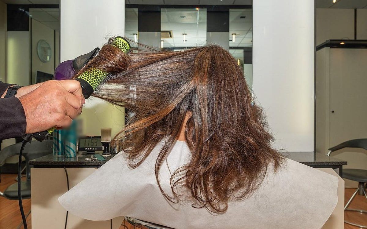 La piega con la spazzola tonda e il phon viola su capelli castani lunghi; la cliente è di spalle.