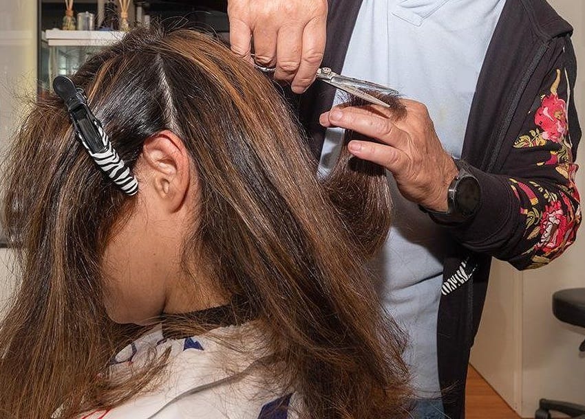 Le mani di Marcello col pettine e le forbici su una ciocca castana; la cliente è di spalle, con una pinza zebrata fra i capelli.