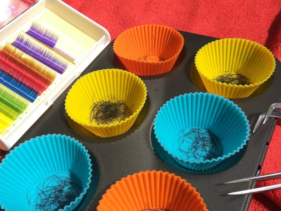 Il loro vassoio delle ciglia: una scatola di extension colorate, viola, rosse, blu, verdi e gialle, le coppette di silicone arancio, gialle e azzurre e la pinzetta, sull'asciugamano rosso.