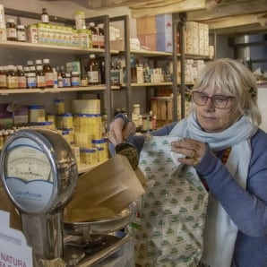 La titolare al banco mentre pesa le erbe sulla bilancia meccanica e riempie un sacchetto di carta