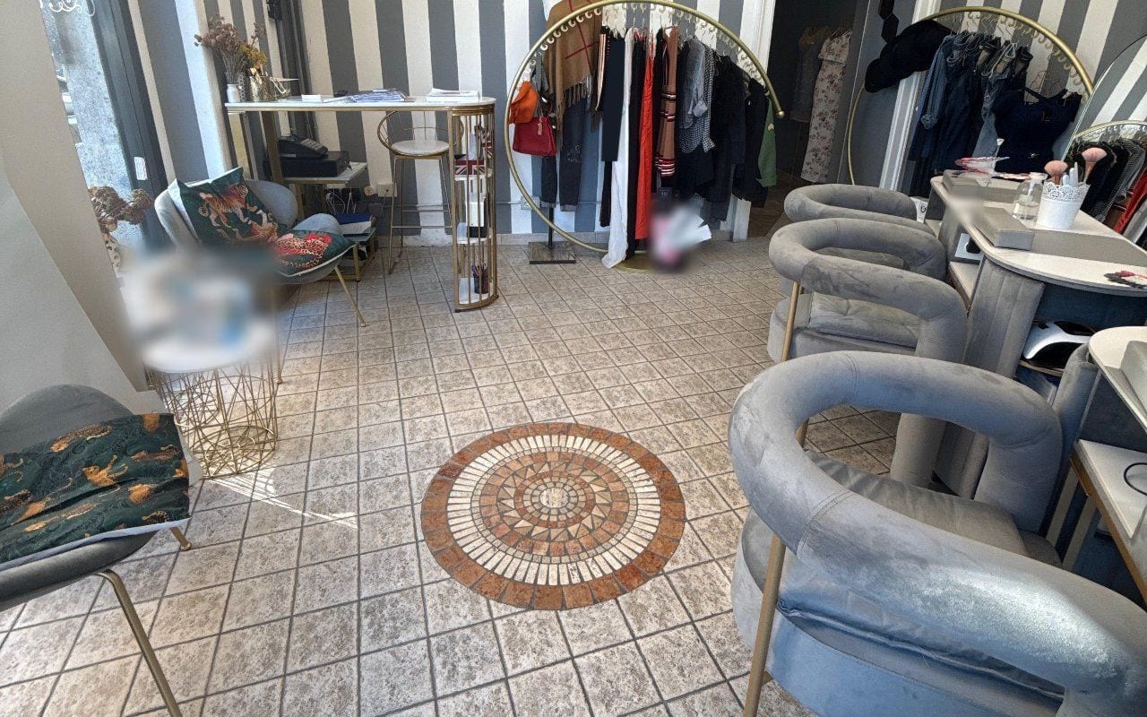 Il salone: pareti a righe grigie e bianche, due stender ad arco dorati con abiti e borse, poltroncine di velluto grigio coi piedi dorati e un rosone di mosaico sul pavimento.