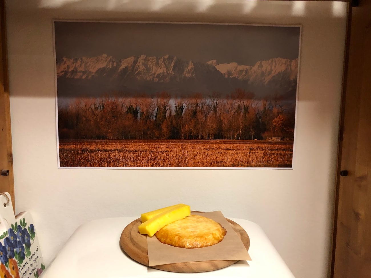 Frico e polenta su un tagliere, sotto la fotografia incorniciata della pianura friulana