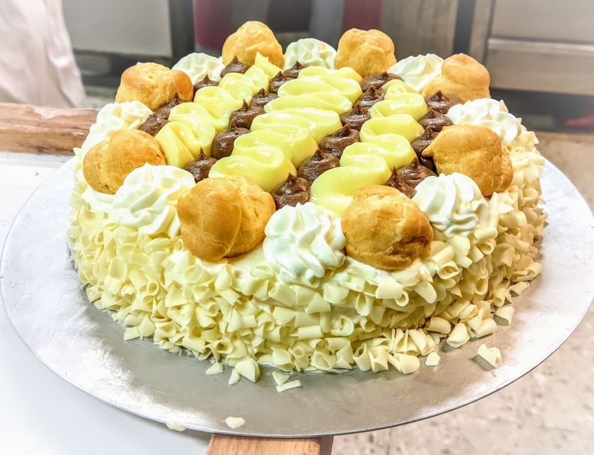 Una torta decorata coi bignè, i ciuffi di panna, la crema gialla e al cioccolato, e i riccioli di cioccolato bianco.