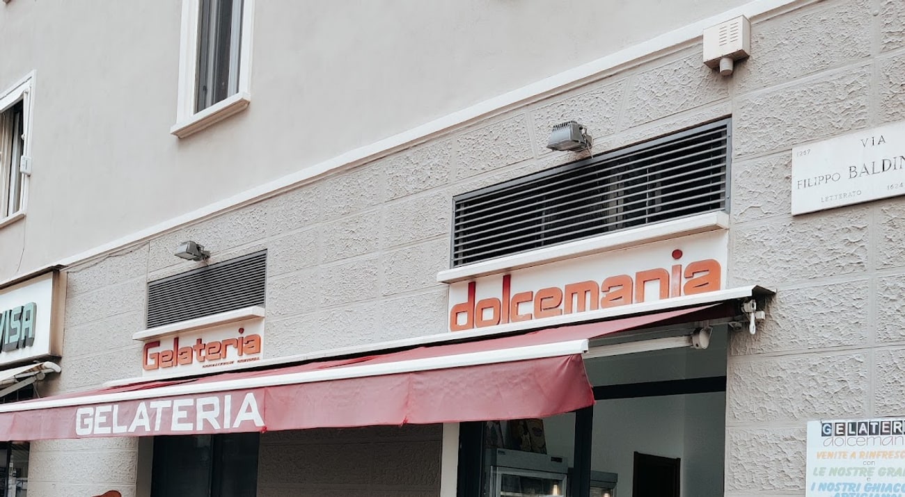 La facciata d'angolo di Dolcemania su Via Baldinucci: insegna arancione, tenda bordeaux e il cono gigante