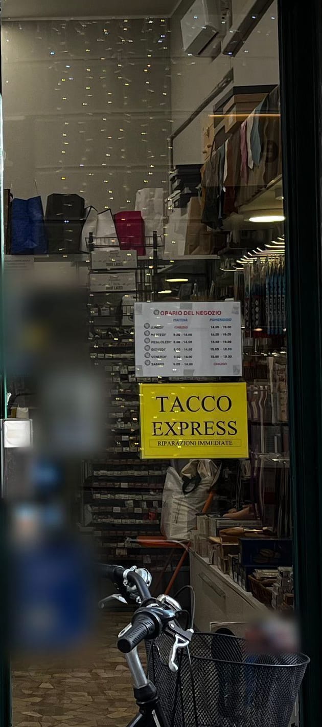 La porta di destra: il cartello «Orario del negozio», il cartello giallo «Tacco Express – riparazioni immediate» e dietro la parete dei cassettini.