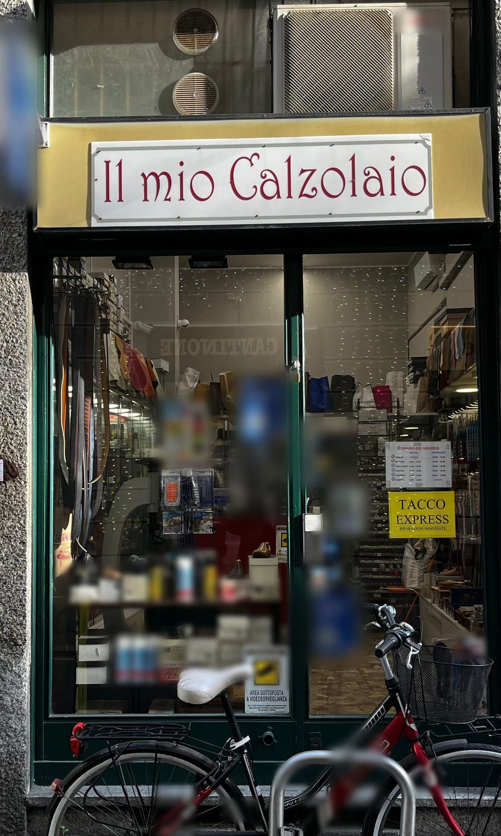 La vetrina di Via dell'Unione 3: l'insegna «Il mio Calzolaio» bordeaux su lastra bianca nella fascia d'oro, il telaio verde, dentro le cinture, i lucidi e il cartello giallo «Tacco Express»; davanti una bicicletta.