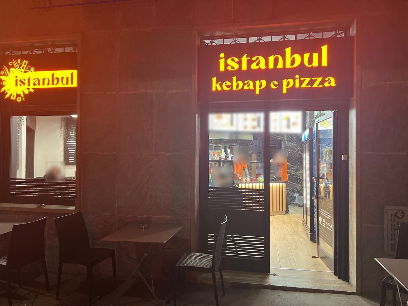 Il locale di sera: l'insegna con la scritta istanbul kebap e pizza accesa gialla sui pannelli neri, la vetrina con lo schizzo di Istanbul, la porta aperta sul banco, i tavolini fuori.