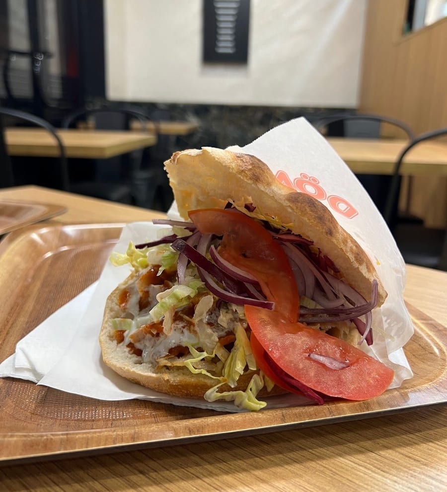 Un panino kebap nel pane tondo, con pomodoro, cipolla rossa e insalata, nella carta su un vassoio di legno.