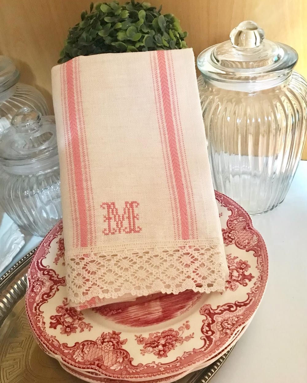 Un asciugapiatti di lino a righe rosa con la M ricamata e il bordo di pizzo, sui piatti di toile rossa.