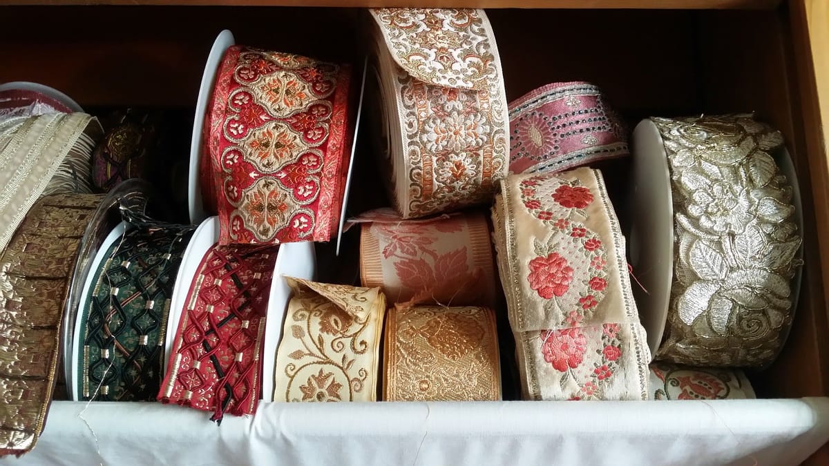 Rotoli di passamanerie jacquard a fiori, rossi, rosa, écru e oro, in un cassetto.