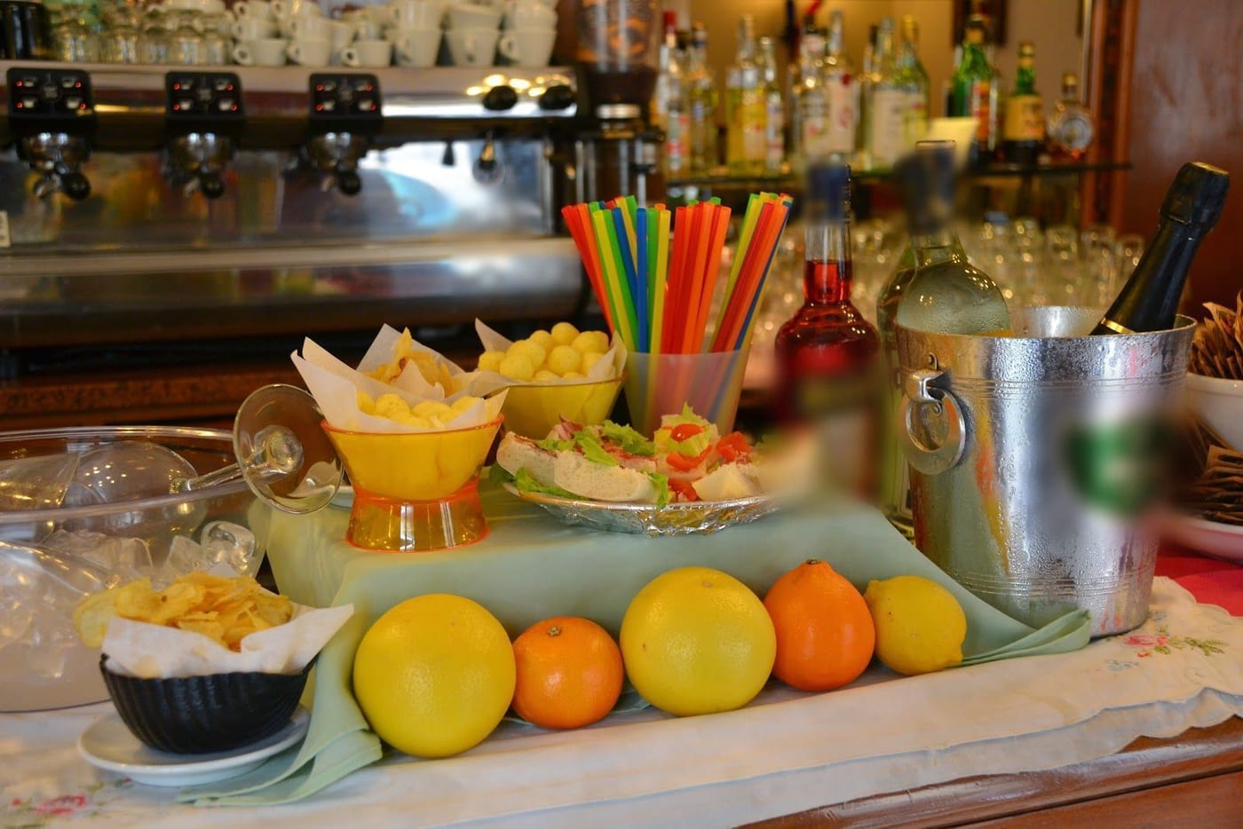 Il banco dell'aperitivo: patatine, tartine, cannucce colorate, bottiglie nel secchiello, pompelmi e arance