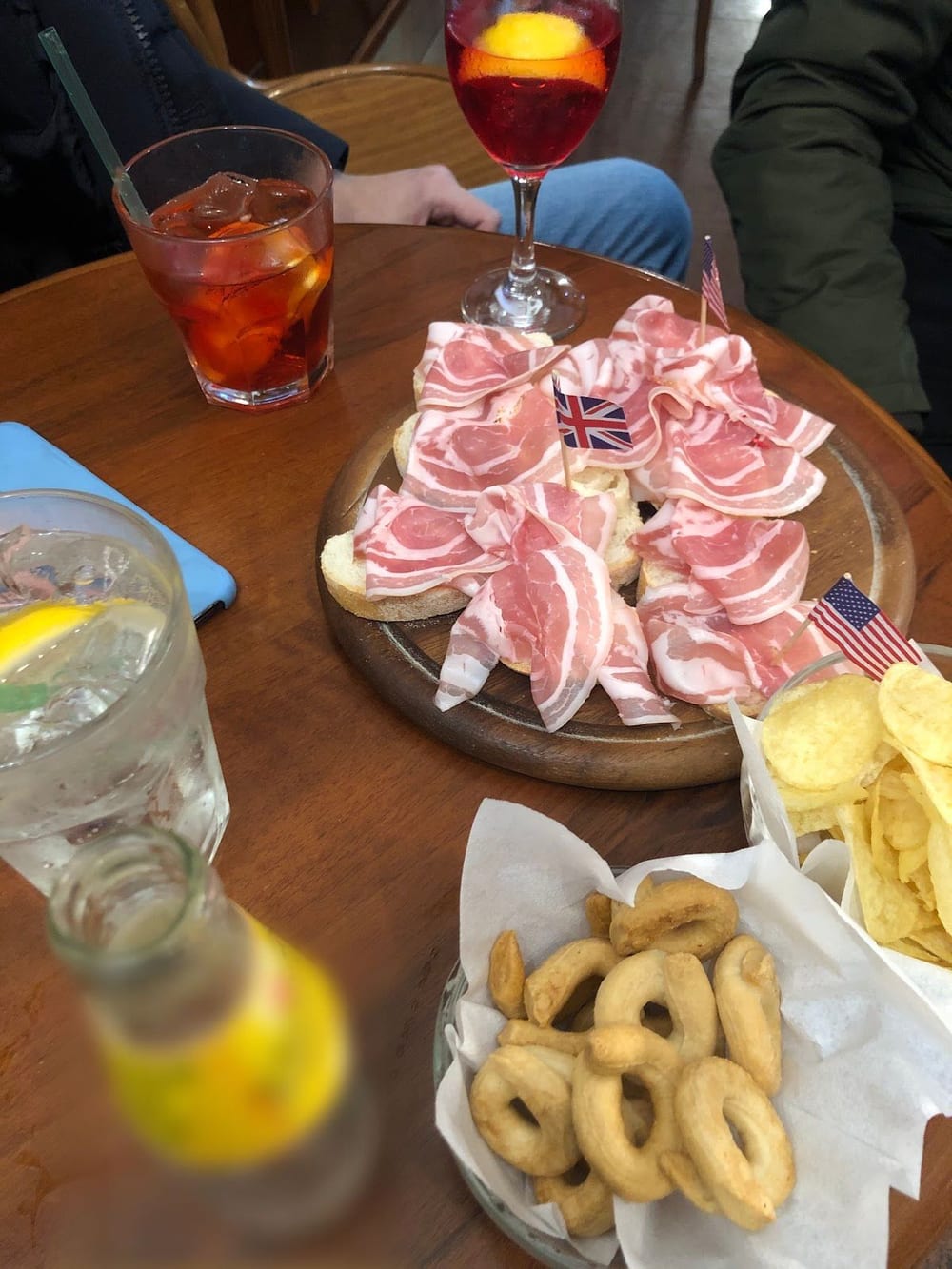 Sul tavolino: due aperitivi, un tagliere di pane e affettato con due bandierine, patatine e taralli