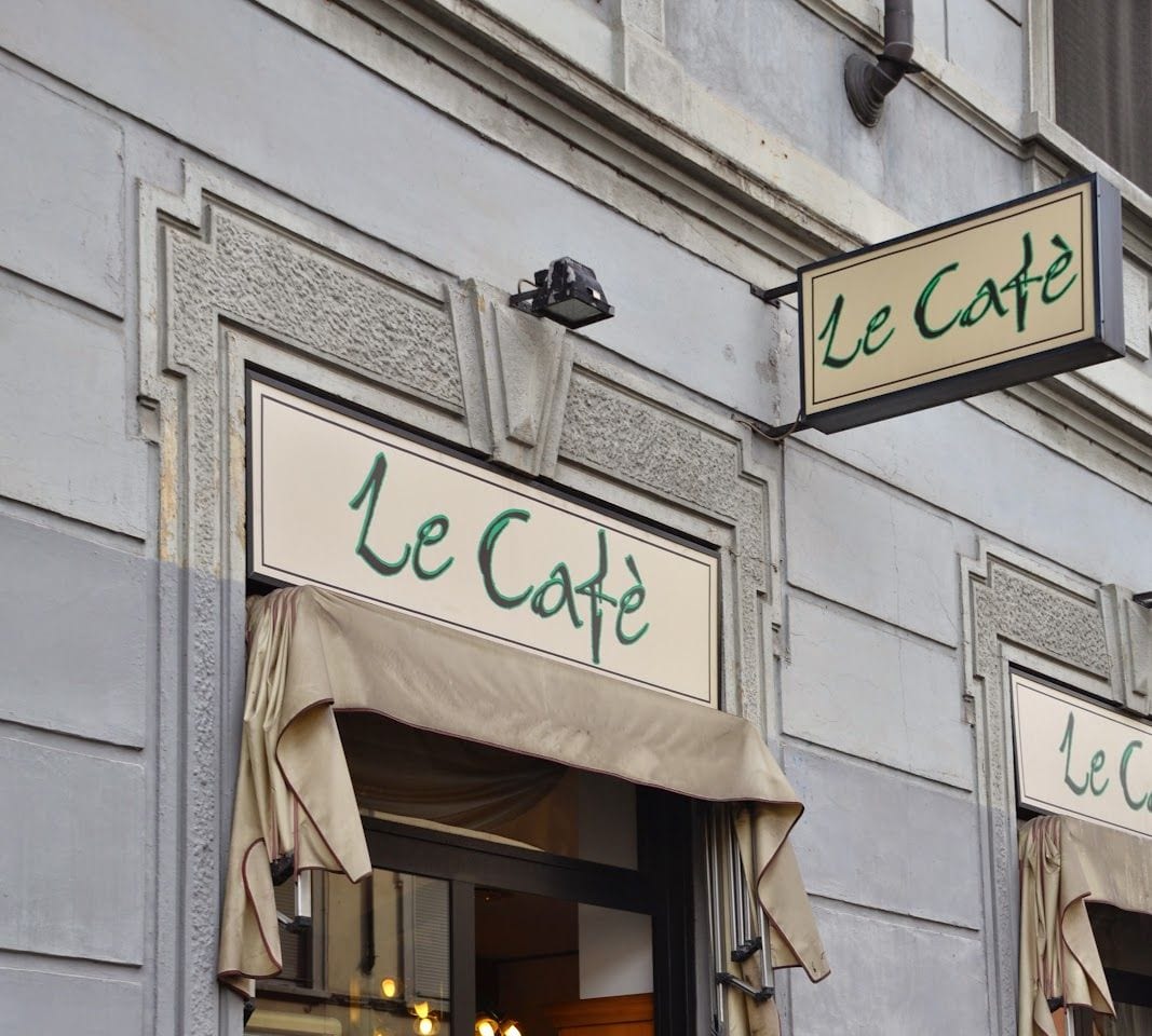 La facciata: l'insegna «Le Cafè» sopra la tenda color sabbia e quella a bandiera, sul palazzo grigio