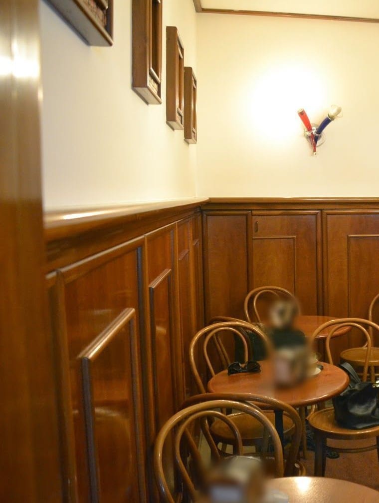 La sala: la boiserie di legno, le cornici alle pareti, una lampada colorata, le sedie di legno curvato attorno ai tavolini rotondi