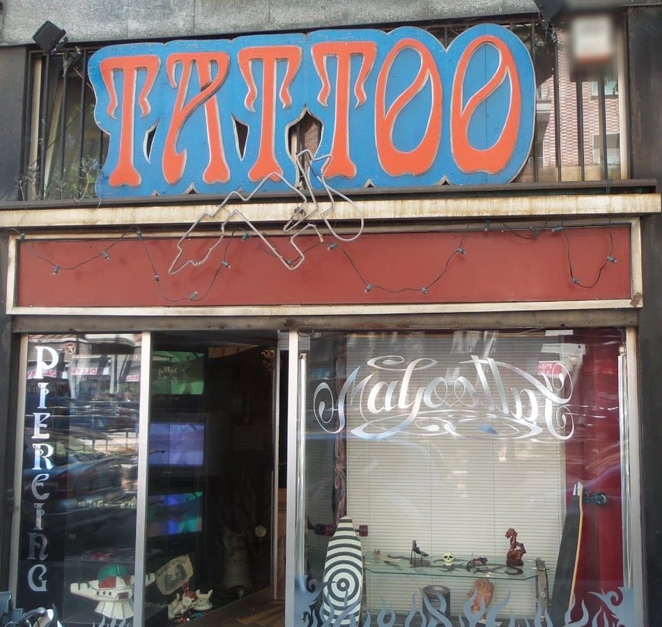 La facciata: l'insegna TATTOO con le lettere arancio su un pannello azzurro, una macchinetta da tatuaggio di fil di ferro appesa sotto, la vetrina con la scritta Magoo Tattoo in corsivo e le fiamme, la scritta PIERCING in verticale sulla porta.