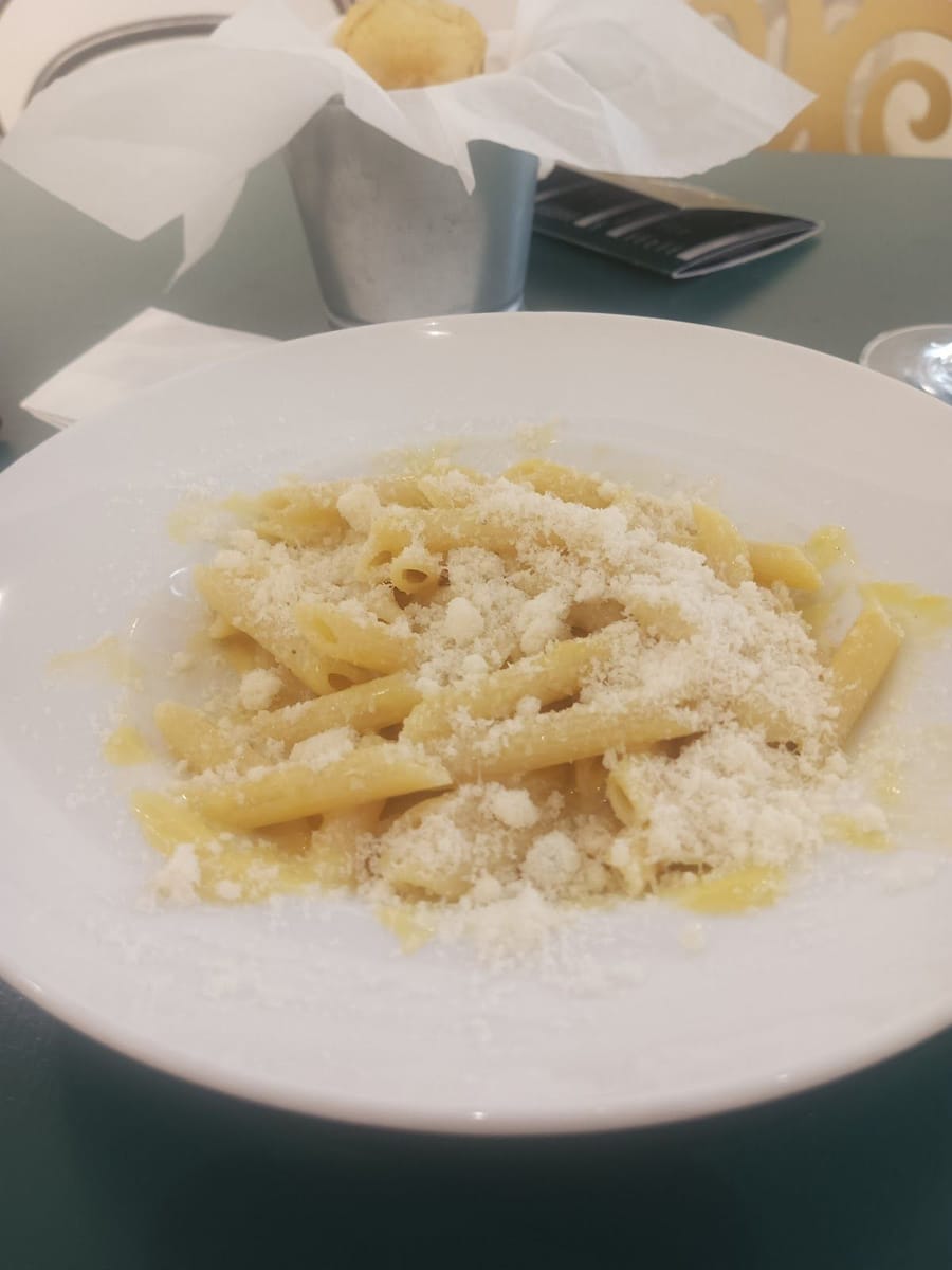 Un piatto di penne col formaggio grattugiato sul tavolino verde, accanto il cestino del pane.