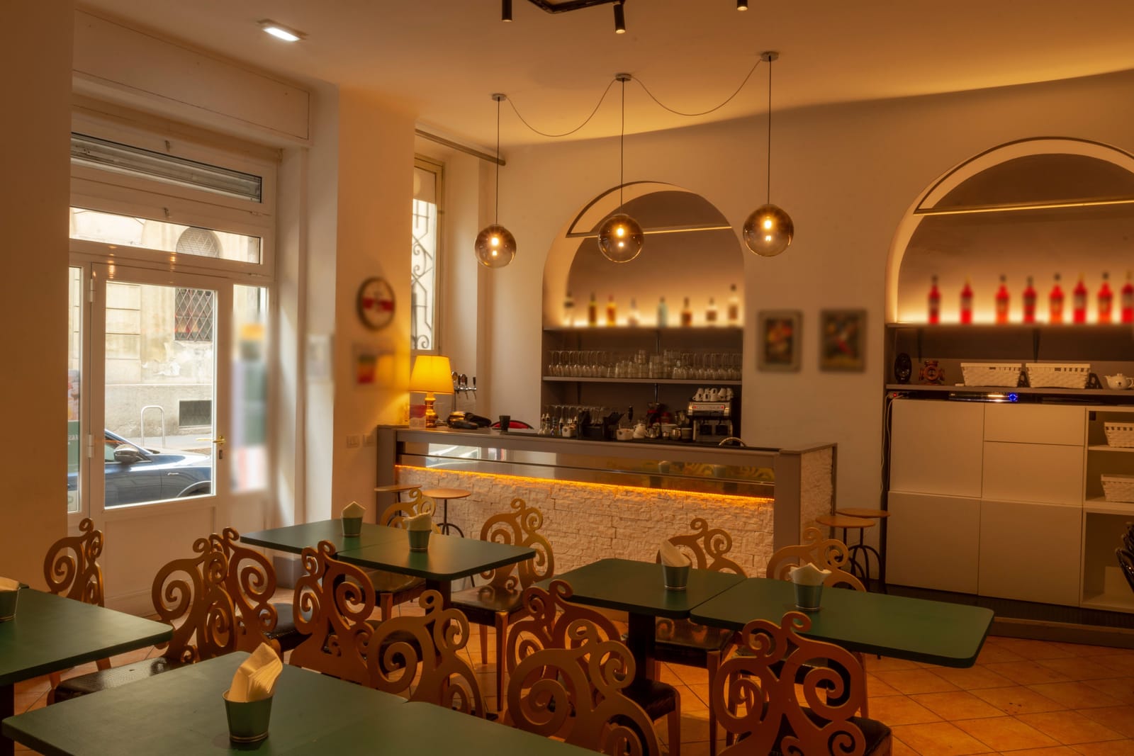 La sala: i tavolini verdi, le sedie dorate a riccioli, le lampade a sfera, le due nicchie ad arco e il bancone di pietra bianca con la luce calda.