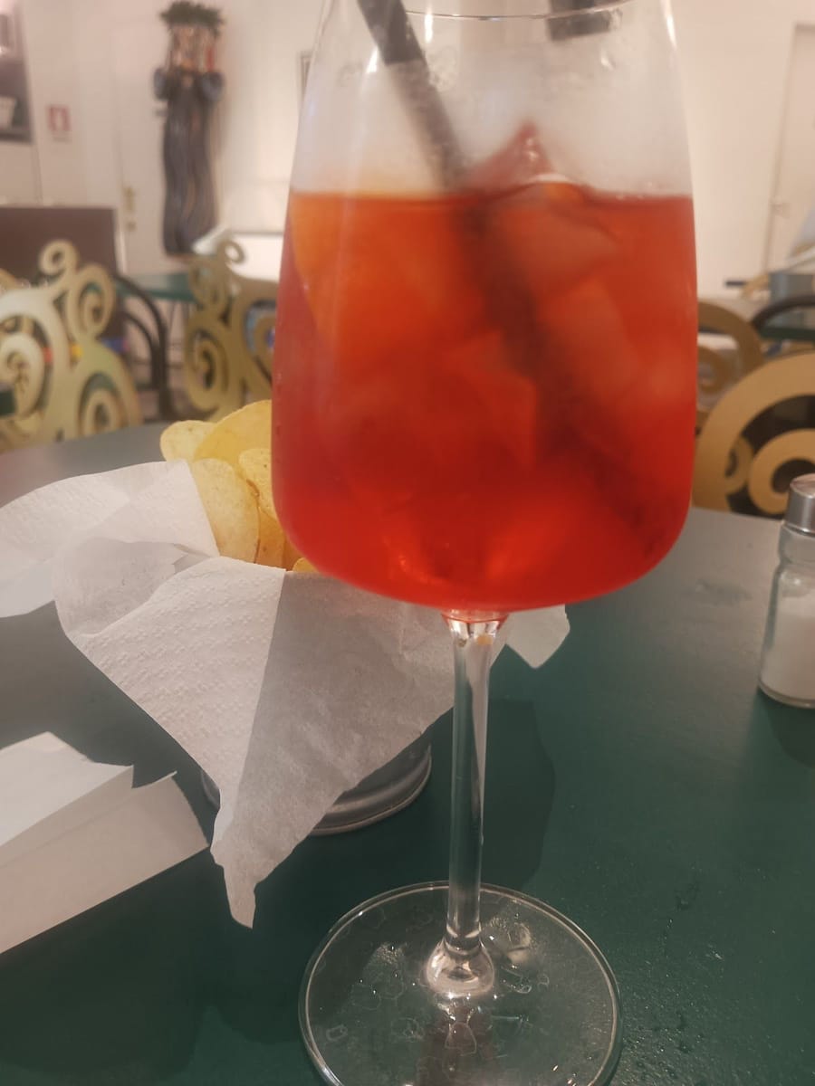 Lo spritz nel calice con la cannuccia sul tavolino verde, dietro le patatine e le sedie dorate.