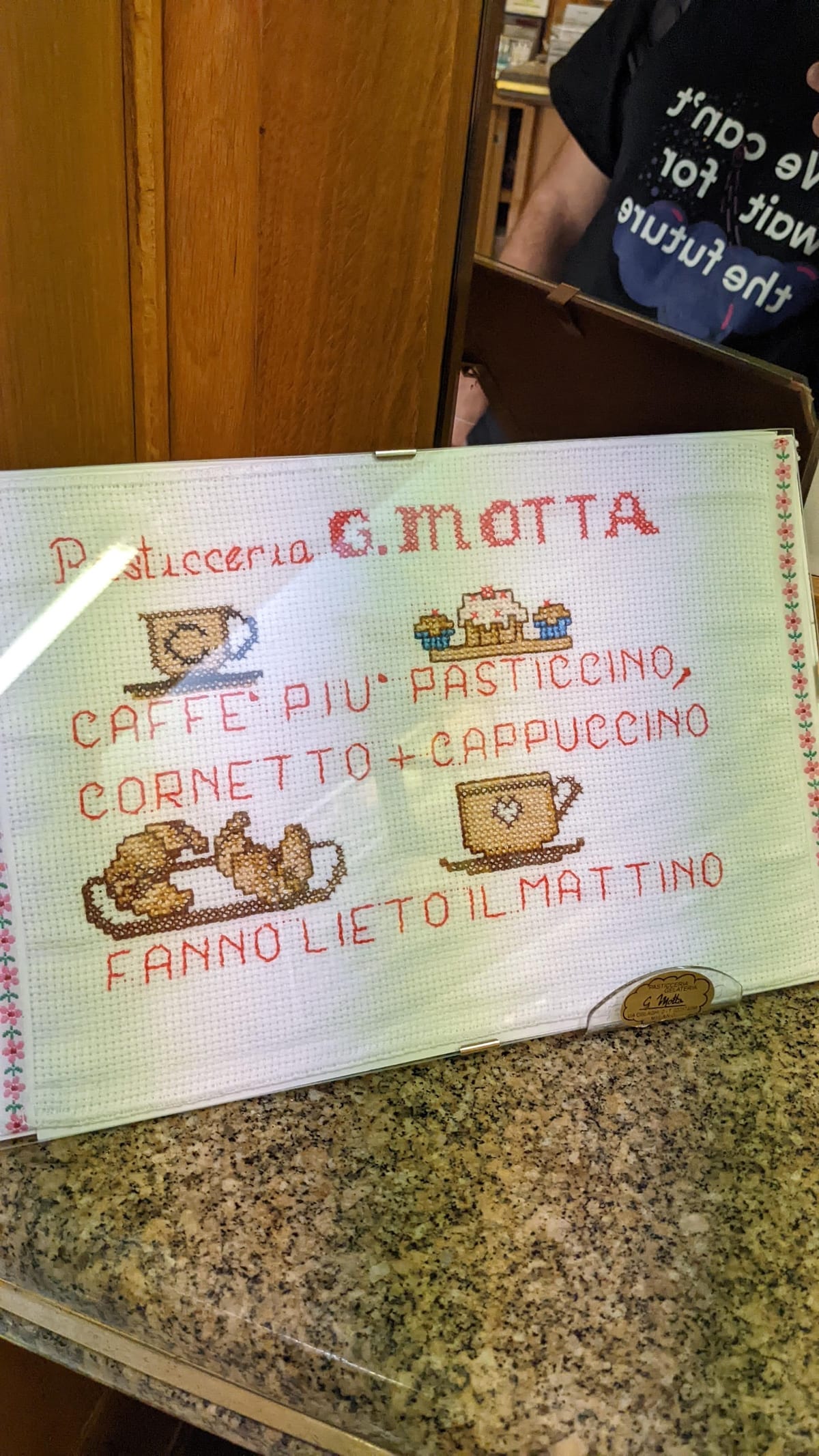 Il ricamo a punto croce incorniciato sul bancone: Pasticceria G. Motta, caffè più pasticcino, cornetto più cappuccino fanno lieto il mattino