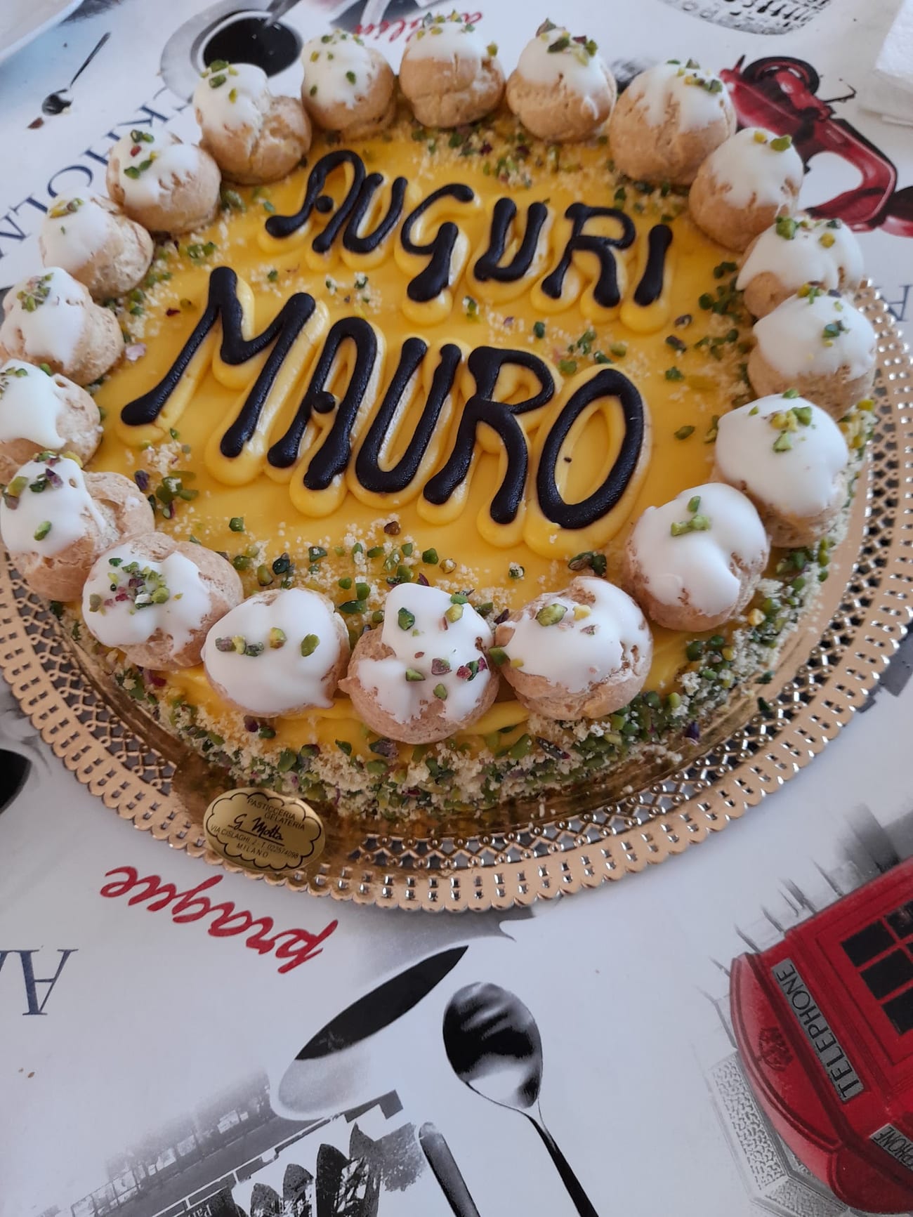 Torta di compleanno alla crema di pistacchio con scritta in cioccolato e corona di bignè, sul vassoio con l'etichetta G. Motta