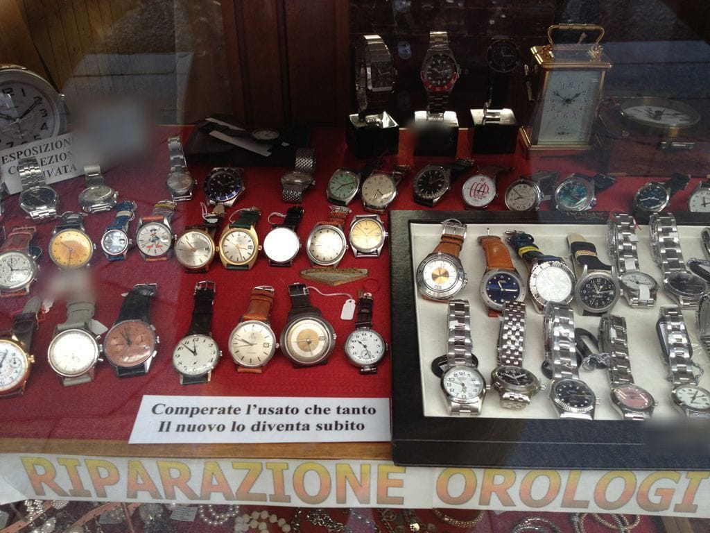 La vetrina piena di orologi da polso sul velluto rosso, una sveglia da viaggio, il cartello «Riparazione orologi».